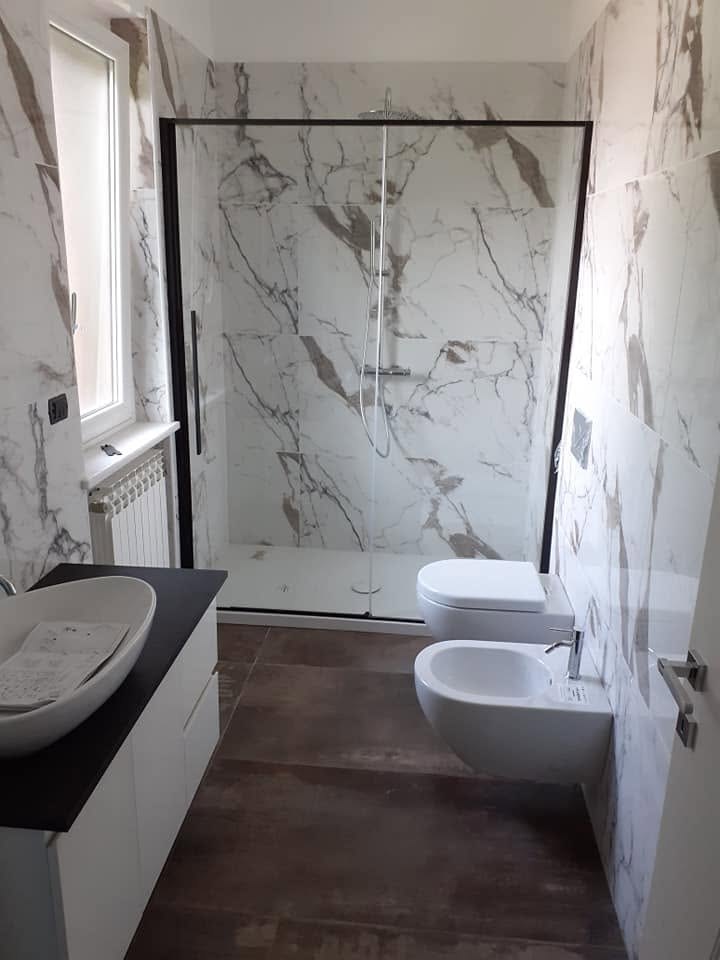Bagno con piastrelle effetto marmo bianco venato, box doccia con profili neri, sanitari sospesi e lavabo d'appoggio su mobile bianco