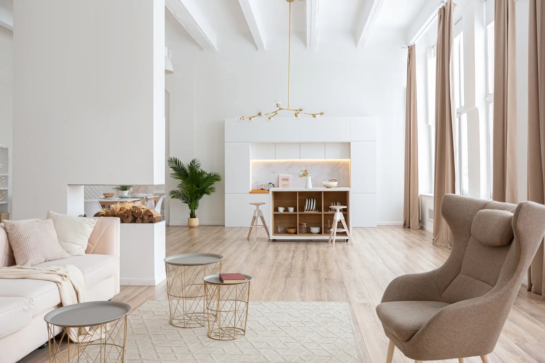 Open space bianco con divano panna, poltrona tortora, tavolini in metallo dorato, cucina con isola e parquet chiaro