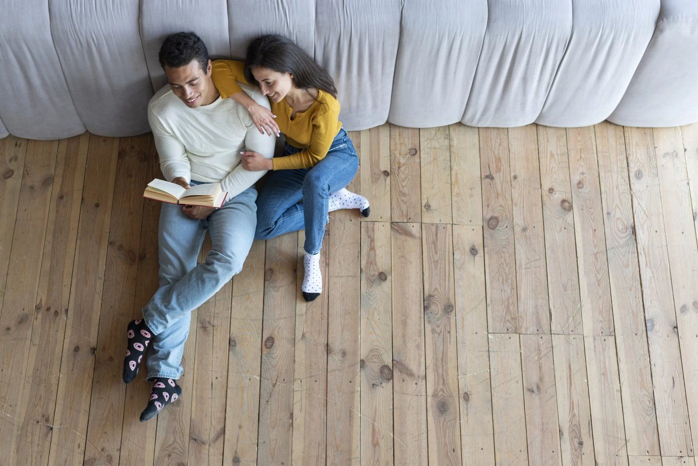 Coppia seduta sul parquet chiaro a listoni contro il divano grigio: lui legge un libro, lei lo abbraccia sorridendo